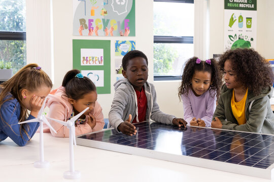 Diverse school children exploring solar panel and wind turbine models in classroom