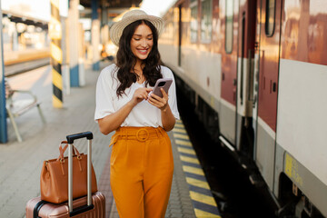 Young attractive lady traveler standing with suitcase at railway station platform with cellphone in...