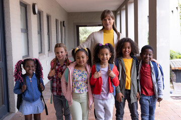Smiling female teacher with diverse group of children wearing backpacks at school