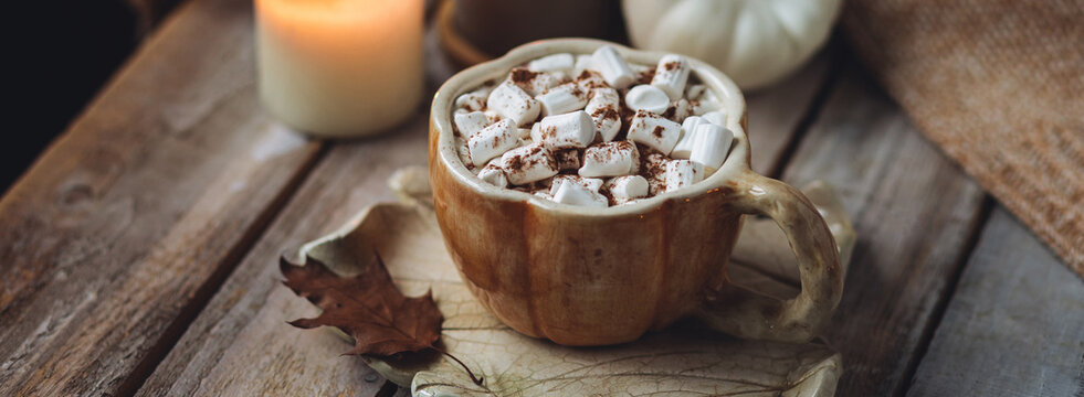 Spicy sweet fall hot drink: delicious pumpkin latte with cinnamon, marshmallow with salted caramel. Served in handmade artisan mug in shape of pumpkins, cozy home decor with candles, dry autumn leaves