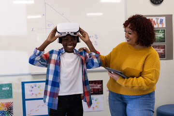 African american boy wearing vr headset in classroom, female teacher holding tablet, at school
