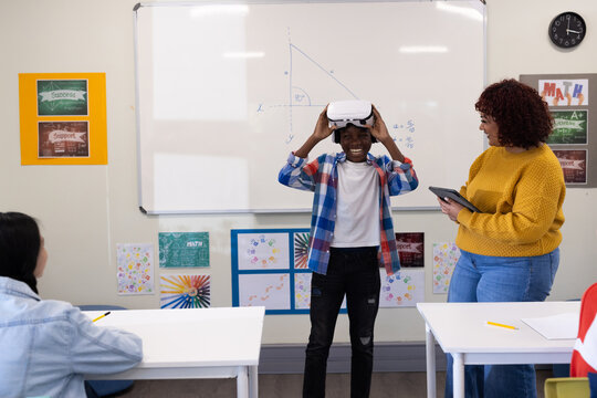 African american boy using vr headset in classroom while female teacher observes, at school