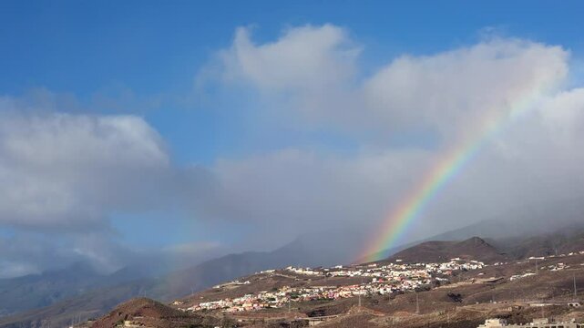 Afterstorm rainbow glows above Tabaiba coastline with dramatic clouds and village nestled in volcanic terrain.