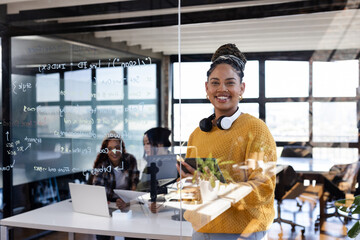 Holding tablet in office, confident woman smiling as colleagues collaborate on laptops