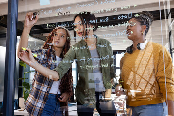 Diverse women collaborating on coding project, writing on glass board in modern office