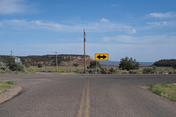 Desert road intersection under a clear blue sky, featuring a yellow directional sign with black...