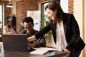 Black man sharing a laughter with his caucasian female coworker at the office desk while having a business discussion. Cheerful multiethnic colleagues having a meeting, reviewing reports on laptop.