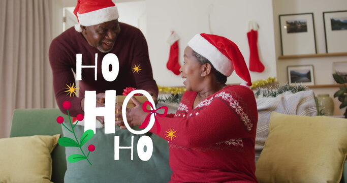 Santa-hatted man giving ribbon-tied gift to woman on sofa in living room, with stockings  and  tinse