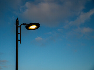 A street LED light is lit up in the evening sky. The sky is mostly clear with a few clouds scattered around. The light is shining brightly, illuminating the area below. The scene has a peaceful mood