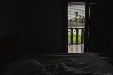 Dark moody bedroom interior with unmade bed and open blinds revealing a scenic terrace and greenery...