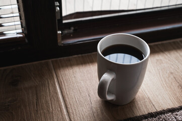 Minimalistic morning scene with a cup of black coffee on a wooden floor near a window, soft sunlight and shadows creating cozy, peaceful atmosphere and warm tones