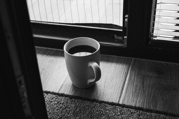 Minimalistic morning scene with a cup of black coffee on a wooden floor near a window, soft sunlight and shadows creating cozy, peaceful atmosphere and warm tones