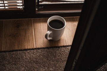 Minimalistic morning scene with a cup of black coffee on a wooden floor near a window, soft sunlight and shadows creating cozy, peaceful atmosphere and warm tones