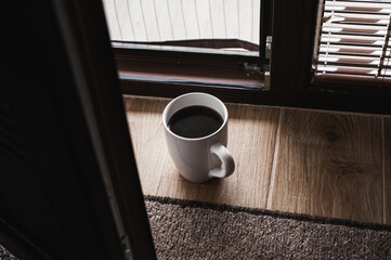 Minimalistic morning scene with a cup of black coffee on a wooden floor near a window, soft sunlight and shadows creating cozy, peaceful atmosphere and warm tones