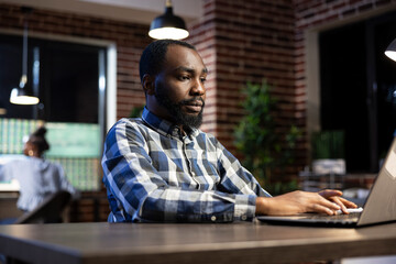 Male financial analyst works late at a brick wall office desk, reviewing documents and stock data...