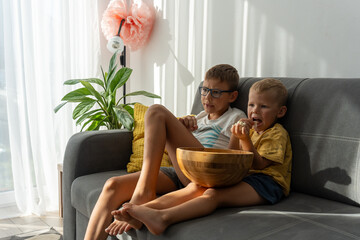 Cute little boys relaxing on soft grey couch in bright living room, sharing large wooden bowl of popcorn and watching TV with full attention. Concept of family time, cozy moments and childhood routine