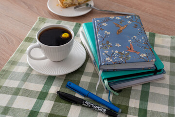 Cozy workspace with a cup of coffee, notebooks, and pens on a green checkered tablecloth, perfect for writing, journaling, or creative work on a calm afternoon. Concept of relaxation and creativity. 