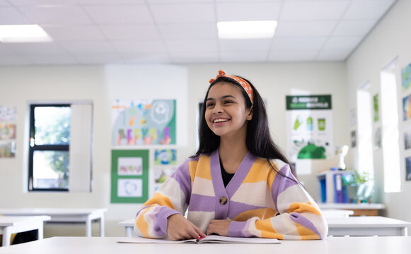 Schoolgirl sitting at white desk reading open book in classroom at school with colorful posters