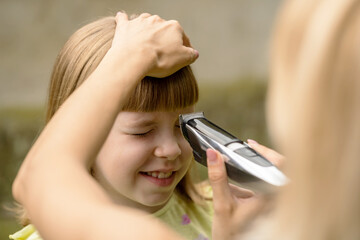 Mother cutting her smiling daughters hair with an electric trimmer outdoors, closeup of a child getting fringe trimmed by a parent, frugal family haircut, bonding moment simple lifestyle concept