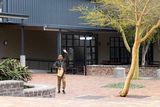 African American boy waving hand and holding brown paper bag near succulent planter in courtyard