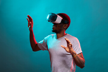 African american adult wearing a VR headset and with raised arms, exploring a dynamic 3D environment. Black male individual standing over blue background, interacting with a virtual interface.