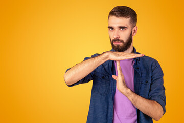 Millennial bearded guy showing TIME OUT or STOP gesture over orange studio background. Young...