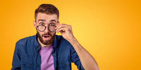 Shocked young guy in glasses opening mouth in disbelief on orange studio background. Overwhelmed...