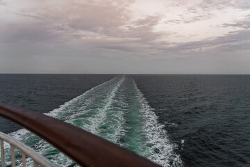 A tranquil view of the wide ocean as a cruise ship creates a wake, leaving behind ripples on the water. The sky carries soft hues of twilight, adding to the serene atmosphere