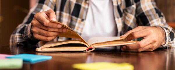 Student Guy Reading Book Learning In Library Sitting At Desk Doing University Homework Indoor. Modern Higher Education And Knowledge Concept. Cropped, Selective Focus