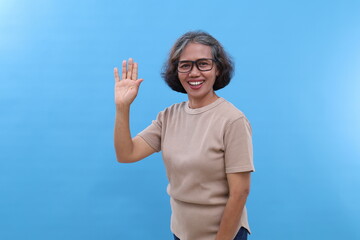 Fototapeta premium Gray-haired Asian old woman, wearing glasses, waving hand, greeting someone in a friendly manner, looking at camera with smile, standing isolated on blue background.