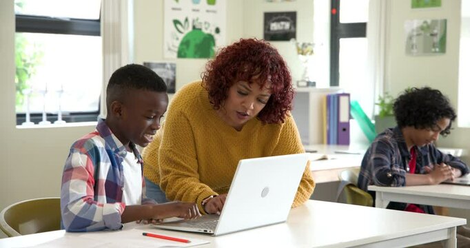 Female teacher helping african american boy with laptop in classroom, focusing on learning, at schoo