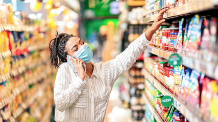 Black Lady Talking On Phone Buying Food In Supermarket. African Woman Chatting On Cellphone Doing Grocery Shopping With Shop Cart Purchasing Food Products In Groceries Store.