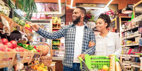 Grocery Shopping In Supermarket. African American Spouses Buying Food And Fresh Fruits Walking With Cart Full Of Products In Shop. Happy Family Buys Groceries Choosing Natural Food In Store Indoors