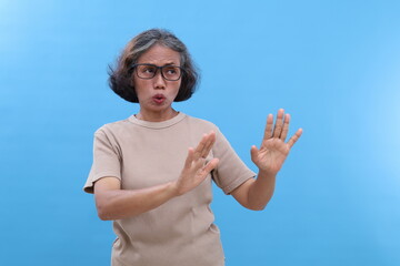 Gray hair Asian old woman, standing looking sideways, wearing glasses, hand gesture of rejection, body facing sideways, dislike expression, isolated blue background.
