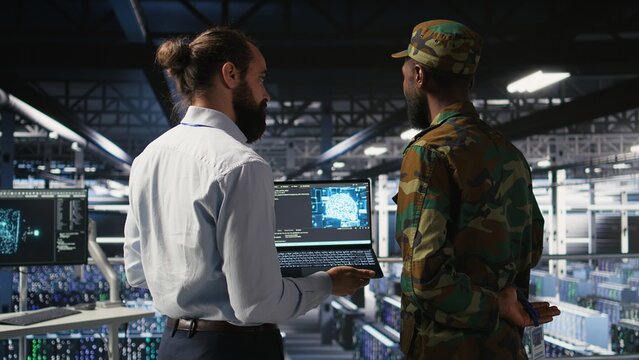 Military technicians in secure data center use laptop to monitor artificial intelligence systems. Army operators in server hub ensuring stable infrastructure for machine learning operations