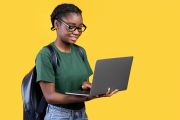 Cheerful Black Student Woman Using Laptop Standing Over Yellow Studio Background, Side View. Remote Education, E-Learning And Online Study Concept. Panorama With Copy Space