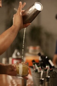 A arte de misturar: um bartender prepara um drink em movimento, com a bebida sendo derramada de uma coqueteleira. A imagem capta a precis&atilde;o e a eleg&acirc;ncia do momento, com um fundo borrado e alegre