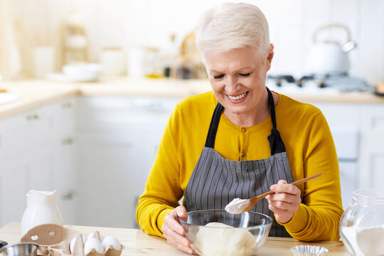 Joyful grandmother wearing apron making dough at kitchen, adding flour, copy space. Happy elderly lady baking pastry cookies or cake at home. Domestic lifestyle, culinary hobby concept