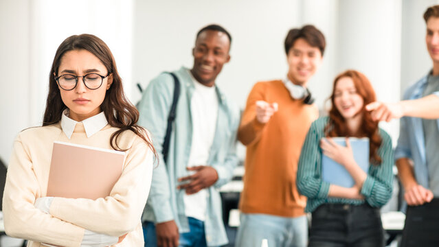 Group Conflict. Sad latin woman in eyeglasses standing away alone, suffering humiliation and public disgrace, crowd of bad people bullying pointing fingers at victim, laughing. Discrimination, Gossip