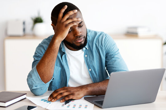 Fototapeta Lack Of Inspiration. Stressed Black Freelancer Guy Sitting At Desk With Laptop And Touching Head In Despair, Having Problems With Project While Working Remotely At Home, Looking At Computer Screen
