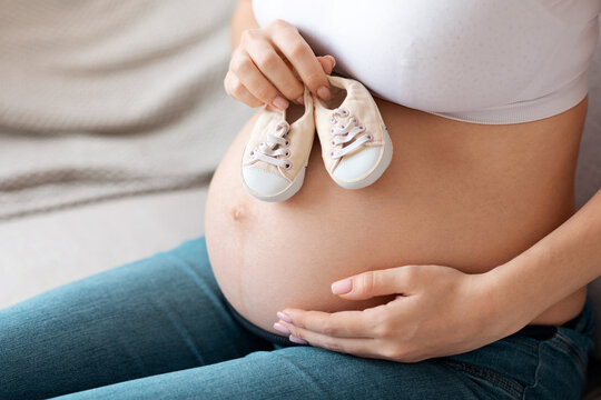 Awaiting Baby. Unrecognizable Pregnant Woman Embracing Tummy And Holding Small Shoes Near Her Naked Belly, Expectant Future Mother Enjoying Pregnancy Time And Maternity Time, Cropped Image, Free Space