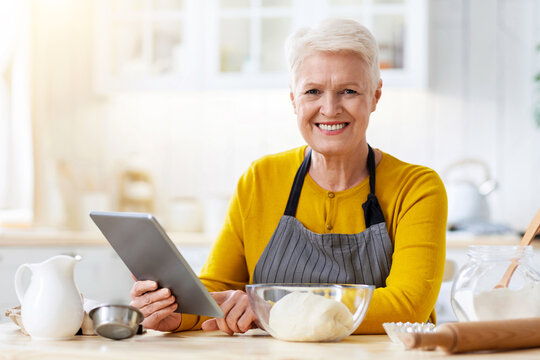 Smiling aged lady cooking in kitchen, looking at digital tablet, watching online culinary class while kneading dough, copy space. Happy old woman cooking by recipes from Internet, using pad