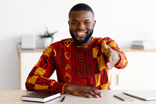 Handsome Black Man In Traditional African Costume Giving Hand For Handshake While Sitting At Desk In Office, Friendly HR Manager Offering Partnership And Job Opportunities, Free Space