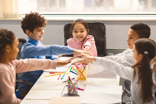 Youth Collaboration Concept. Group of multiethnic elementary schoolchildren sitting in circle at table and putting hands together in stack, having fun. Friends and fellowship, partnership and support - Powered by Adobe