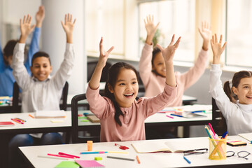 Portrait of multiciltural excited diverse group of small happy elementary school children sitting at desks in classroom, raising hands high up and waving, having fun, learning with pleasure