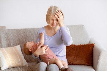 Exhausted blonde woman young mother having migraine, holding crying little baby, home interior,...