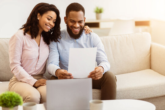 Portrait of cheerful african american couple sitting on the couch in living room, man holding paper, working and reading letter with good news at home with his wife who embracing him - Powered by Adobe