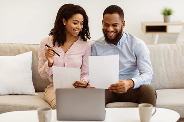 Planning Family Budget. Smiling black couple holding papers, calculating domestic bills and checking household expenses, reading documents or receipt from bank, talking about investment and finance