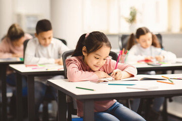 Education And Studying Concept. Portrait of smiling asian small student sitting at desk in classroom, writing in notebook or drawing picture with colorful pencils. Back To School