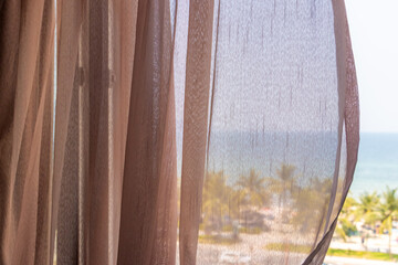 Soft transparent curtains gently blowing beside a window with a tropical view of the sea beach with palm trees